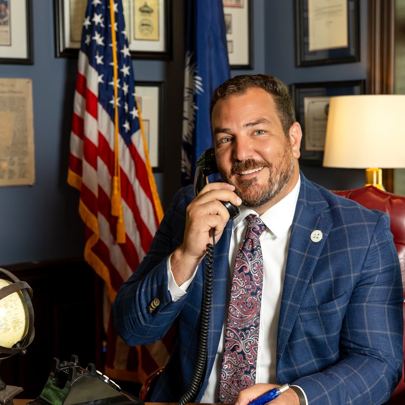 Gabriel Penfield smiling on the phone at his desk, with U.S. and South Carolina flags behind him